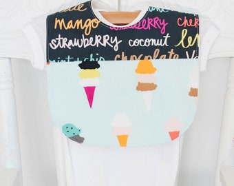 May include: A baby bib with a black top section featuring colorful text of ice cream flavors like mango and strawberry. The bottom section is light blue with ice cream cone illustrations. The bib is hanging on a white onesie.