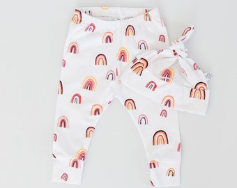 May include: White baby leggings and a matching knotted hat, both adorned with a repeating pattern of colorful rainbows. The rainbows feature shades of red, orange, and yellow. The set is designed for infants and toddlers.