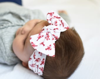 May include: A white headband with a red lobster print tied in a bow. The headband is worn by a baby.