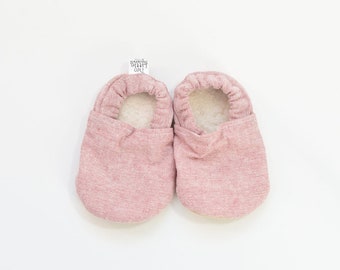 May include: A pair of baby booties in a soft pink hue. The exterior has a textured appearance, while the interior is lined with a fluffy, white material. A small tag with the text "Two Little Beans" is attached to the booties.