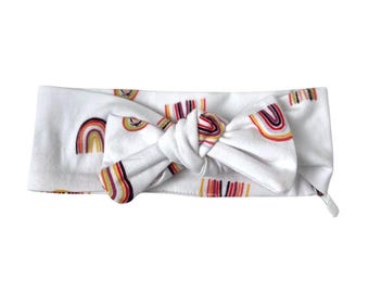 May include: White headband with a knotted bow and a rainbow pattern. The headband is made of soft, stretchy fabric and is perfect for adding a touch of color to any outfit.