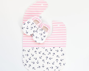 May include: Pink and white striped baby bib with a pocket and a white background with a navy blue anchor pattern. Two white baby booties with a navy blue anchor pattern are on top of the bib.