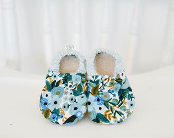 May include: A pair of baby booties with a floral pattern in shades of blue, green, and gold. The booties are made of soft fabric and have a white background.