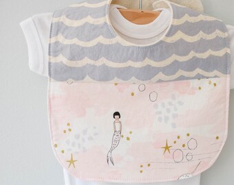 May include: A baby bib with a gray wave pattern at the top and a pink and white watercolor design with a mermaid illustration on the bottom. The bib is hanging on a white onesie.