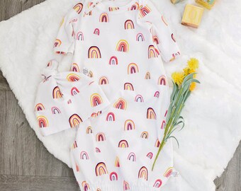 May include: A white baby gown with a rainbow pattern. The gown has long sleeves and a gathered bottom. A matching rainbow hat is also included.