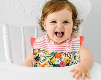 May include: A baby wearing a pink and white striped bib with a floral pattern. The bib has a ruffled edge and is made of a soft, comfortable fabric. The baby is sitting in a white high chair and is smiling.