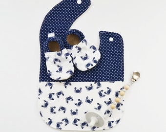 May include: A baby bib, shoes, and pacifier set. The bib is navy blue with white polka dots on the top and white with navy blue crab illustrations on the bottom. The shoes and bib feature a matching crab design. A pacifier and wooden bead clip are also included.