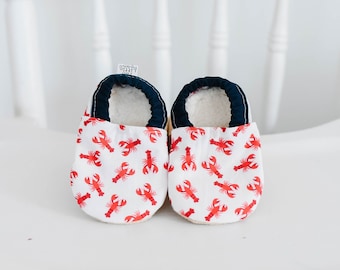 May include: A pair of baby shoes with a white background and a red lobster pattern. The shoes have a navy blue trim and a soft, white interior. The shoes are designed for infants and toddlers.