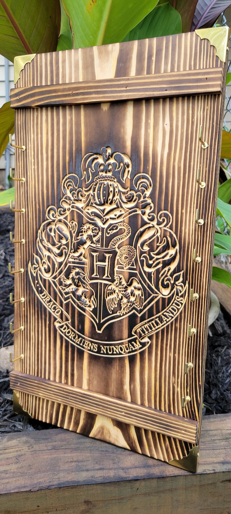 May include: Wooden wall-mounted organizer with a burned finish, featuring the Hogwarts crest. The design includes brass hooks and corner accents. The wood grain is visible, and the piece has a rustic aesthetic.