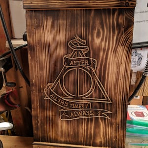 May include: A wooden box with a dark brown, burnt finish. The box features the Deathly Hallows symbol and the text "AFTER", "ALL THIS TIME?", and "ALWAYS" carved into the wood. The corners are reinforced with brass-colored metal.