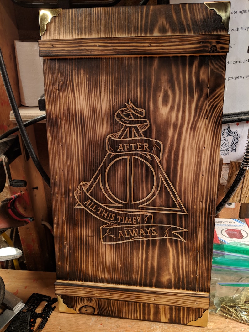 May include: A wooden box with a dark brown, burnt finish. The box features the Deathly Hallows symbol and the text "AFTER", "ALL THIS TIME?", and "ALWAYS" carved into the wood. The corners are reinforced with brass-colored metal.