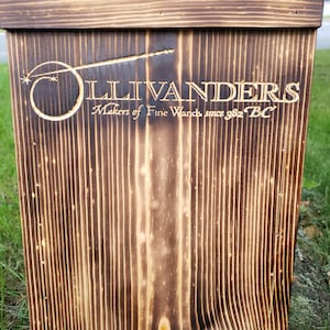 May include: A wooden sign with the text "Ollivanders Makers of Fine Wands since 382 BC" is displayed. The sign is made of dark brown wood with vertical grain lines and a burned finish. The Ollivanders logo is a wand with a star and a circle.