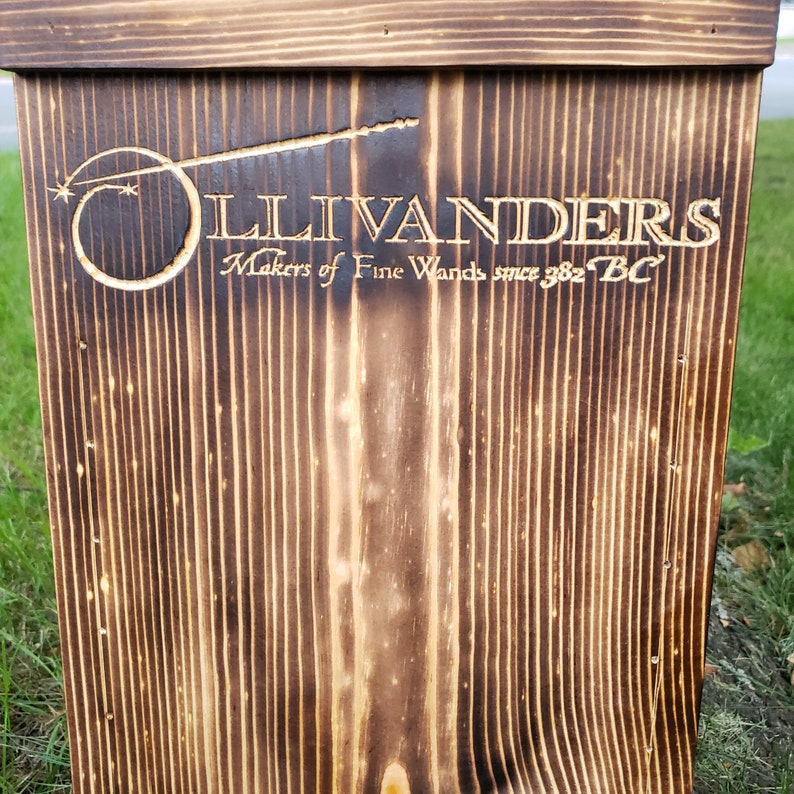 May include: A wooden sign with the text "Ollivanders Makers of Fine Wands since 382 BC" is displayed. The sign is made of dark brown wood with vertical grain lines and a burned finish. The Ollivanders logo is a wand with a star and a circle.