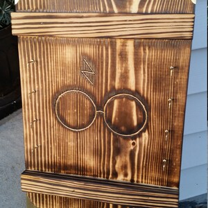 May include: A wooden wall-mounted shelf with a dark brown finish. The shelf features a carved design of round glasses and a lightning bolt. The corners are reinforced with brass-colored metal. The shelf is made of vertical wooden planks.
