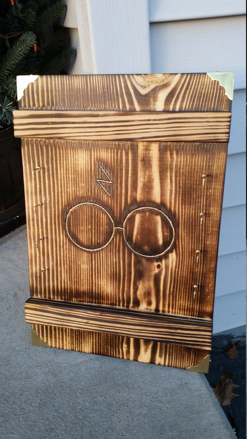 May include: A wooden wall-mounted shelf with a dark brown finish. The shelf features a carved design of round glasses and a lightning bolt. The corners are reinforced with brass-colored metal. The shelf is made of vertical wooden planks.