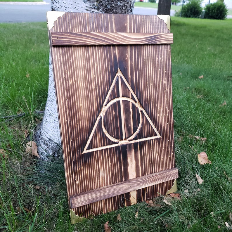 May include: A wooden sign with a dark, burnt finish, featuring the Deathly Hallows symbol. The sign has a rectangular shape with decorative corner accents and a horizontal support piece. The sign is leaning against a tree on a grassy surface.