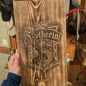 May include: A wooden sign with a dark brown finish and a carved Slytherin crest. The crest features a snake and the word "Slytherin" in a banner. The sign has a rectangular shape with decorative top and bottom edges.