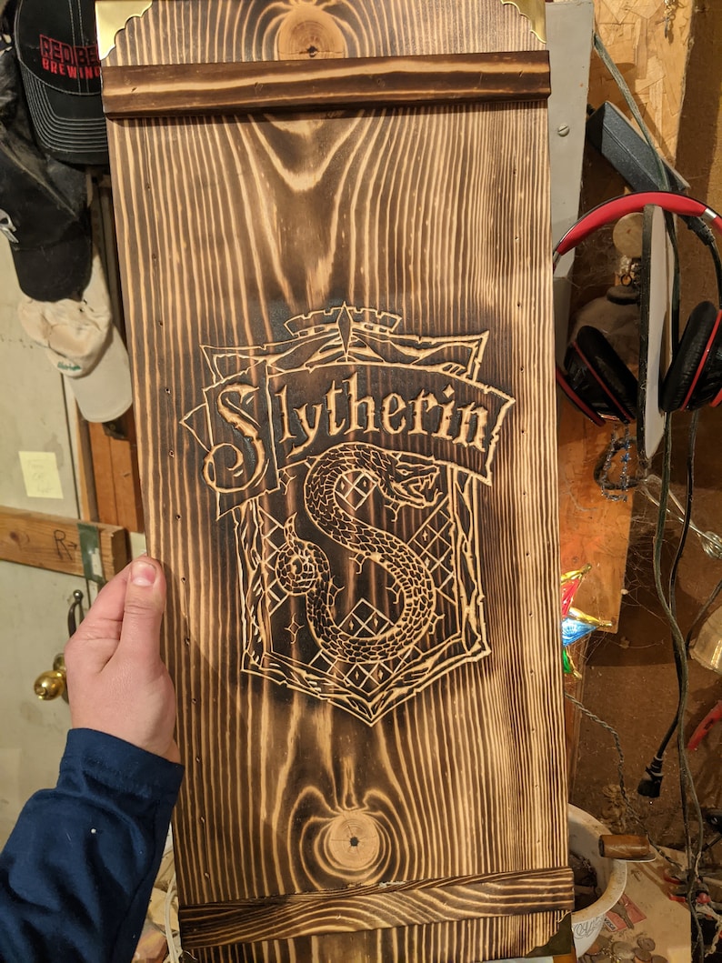 May include: A wooden sign with a dark brown finish and a carved Slytherin crest. The crest features a snake and the word "Slytherin" in a banner. The sign has a rectangular shape with decorative top and bottom edges.