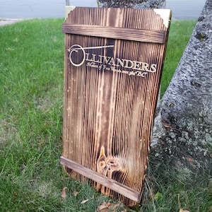 May include: A wooden sign with the text "OLLIVANDERS Makers of Fine Wands since 982 BC" in gold lettering. The sign is made of dark stained wood with brass corner accents and a wooden frame. The sign is leaning against a tree.