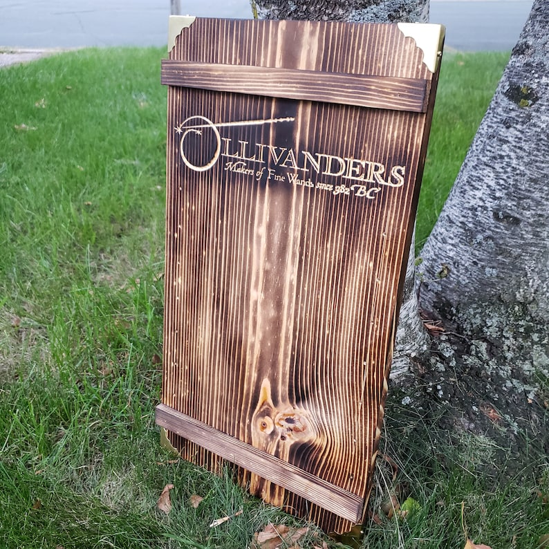 May include: A wooden sign with the text "OLLIVANDERS Makers of Fine Wands since 982 BC" in gold lettering. The sign is made of dark stained wood with brass corner accents and a wooden frame. The sign is leaning against a tree.