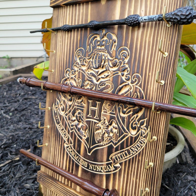 May include: Wooden wand display featuring the Hogwarts crest and the words "Draco Dormiens Nunquam Titillandus". The display has three wands and brass hooks. The wands are dark brown and black.