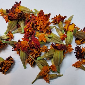 May include: Dried marigold flowers and buds, featuring vibrant orange and deep red petals. The image shows a collection of dried flowers and buds, with green stems and sepals. The flowers are scattered on a white surface, showcasing their natural colors and textures.