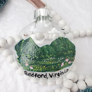 May include: Clear glass Christmas ornament with a hand-painted landscape scene. The scene depicts green mountains, a lake, and wildflowers, with the text "Bedford, Virginia" at the bottom. The ornament has a silver cap and a brown velvet ribbon.