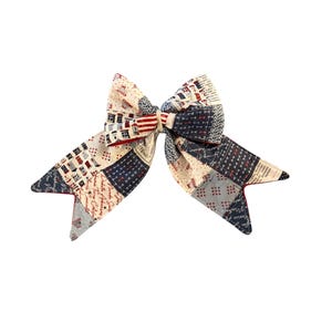 May include: A fabric hair bow featuring a patchwork design with red, white, and blue colors. The pattern includes illustrations of small houses, stars, stripes, and cursive text. The bow has two long, pointed tails and a central knot detail.