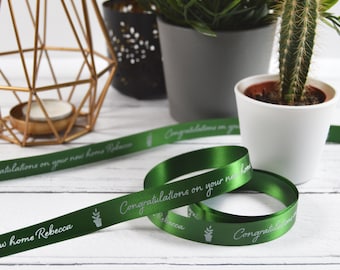 May include: A dark green satin ribbon featuring white printed text that reads Congratulations on your new home Rebecca, accompanied by a small potted plant icon. The ribbon is coiled and laid out flat, displaying the repeating message and graphic.