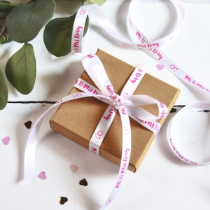 K&ouml;nnte beinhalten: Eine quadratische Geschenkbox aus braunem Karton, die mit einem wei&szlig;en Satinband verschn&uuml;rt ist. Das Band ist mit dem wiederkehrenden rosa Schriftzug Mr & Mrs Day und einem Symbol aus zwei ineinander verschlungenen Ringen bedruckt.