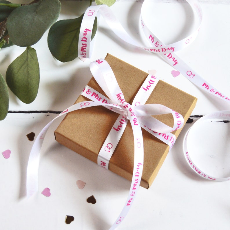 K&ouml;nnte beinhalten: Eine quadratische Geschenkbox aus braunem Karton, die mit einem wei&szlig;en Satinband verschn&uuml;rt ist. Das Band ist mit dem wiederkehrenden rosa Schriftzug Mr & Mrs Day und einem Symbol aus zwei ineinander verschlungenen Ringen bedruckt.