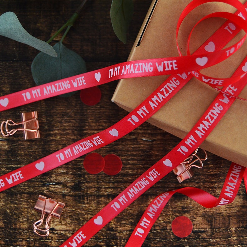 May include: A red satin ribbon featuring the repeating white text TO MY AMAZING WIFE interspersed with small white heart icons. The ribbon is draped across a brown cardboard box, accompanied by several metallic copper-colored binder clips.