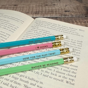 May include: Four hexagonal wooden pencils in pastel shades of teal, pink, light blue, and light green. Each pencil features a gold-coloured metal ferrule, a white eraser, and black text: ONE MORE CHAPTER, I LOVE BOOKS, HERE FOR THE PLOT TWIST, and RATHER BE READING.
