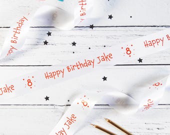 Könnte beinhalten: Ein weißes Satinband mit dem wiederholten roten Text Happy Birthday Jake und der Zahl 8, umgeben von kleinen Sternen. Das Band ist mit verstreuten schwarzen Sternsymbolen verziert und enthält zwei dünne goldene Metallgeburtstagskerzen.
