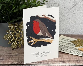 Könnte beinhalten: Eine weiße Grußkarte mit einem Rotkehlchen mit roter Brust, das auf einem schneebedeckten Ast vor einem dunkelblauen Hintergrund mit weißen Schneeflocken sitzt. Der Text Thinking of You AT CHRISTMAS steht in schwarzer Schrift auf der Karte.