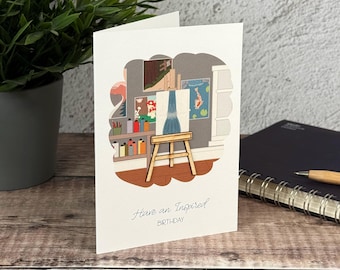 Könnte beinhalten: Eine weiße Grußkarte mit einer zentralen Illustration eines Künstlerateliers mit Staffelei, Leinwand und Malbedarf. Der Text "Have an Inspired BIRTHDAY" ist in blauer Schrift unter der Illustration auf der Vorderseite der Karte gedruckt.