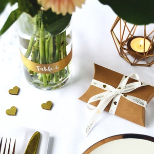 K&ouml;nnte beinhalten: Eine braune Kissenverpackung aus Karton, gebunden mit einem wei&szlig;en Band mit goldenen Herzsymbolen und dem in Schreibschrift gedruckten Namen Aimee. Eine klare Glasvase mit einem goldenen Band mit der Aufschrift Table 6, ein goldener Kerzenhalter.