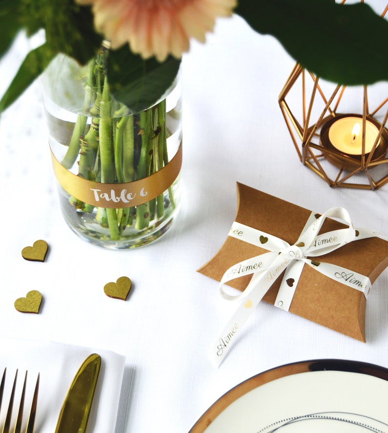 K&ouml;nnte beinhalten: Eine braune Kissenverpackung aus Karton, gebunden mit einem wei&szlig;en Band mit goldenen Herzsymbolen und dem in Schreibschrift gedruckten Namen Aimee. Eine klare Glasvase mit einem goldenen Band mit der Aufschrift Table 6, ein goldener Kerzenhalter.