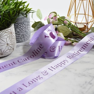 Puede incluir: Cinta de satén morado con el texto "To a Long & Happy Retirement Caroline & H" y un icono de regadera.