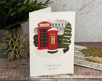 Könnte beinhalten: Eine weiße Grußkarte mit einer roten britischen Telefonzelle und einem roten Briefkasten im Schnee. Ein grüner Weihnachtsbaum steht neben einem schwarzen Zaun. Der Text With love at CHRISTMAS ist in grauer Schrift auf der Vorderseite zu sehen.