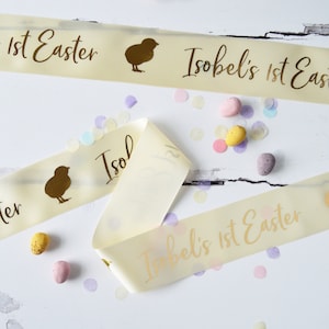 Könnte beinhalten: Ein Set aus drei weißen Satinbändern mit goldener Beschriftung und einer Küken-Silhouette. Die Bänder lauten "Isobel's 1st Easter".