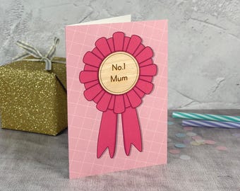Könnte beinhalten: Eine rosa Grußkarte mit einer großen, leuchtend rosa Rosette und einem zentralen Holzkreis. Die Holzmitte ist mit dem Text No.1 Mum graviert. Die Karte hat ein dezentes weißes Gittermuster auf dem rosa Hintergrund, was ein schlichtes Design ergibt.
