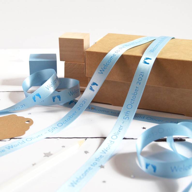 May include: A light blue satin ribbon printed with dark blue baby footprints and the text Welcome to the World Oliver ~ 5th October 2021. The ribbon is draped over a brown cardboard box and coiled near a small blue wooden block and a pencil.