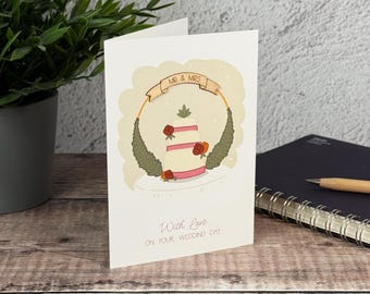 Könnte beinhalten: Eine weiße Grußkarte mit einer Illustration einer dreistöckigen Hochzeitstorte mit rosa Bändern und roten Blumen, umrahmt von grünen Blättern und einem Banner mit der Aufschrift MR & MRS. Darunter steht With Love ON YOUR WEDDING DAY.