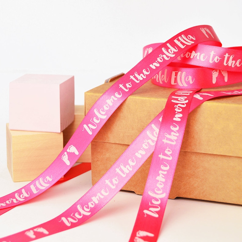 May include: A bright pink satin ribbon featuring the white printed text Welcome to the world Ella and small white baby footprint icons. The ribbon is coiled and draped over a brown cardboard box, with a light pink wooden block visible nearby.