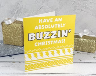 Könnte beinhalten: Eine quadratische gelbe Grußkarte mit weißem Text, der HAVE AN ABSOLUTELY BUZZIN' CHRISTMAS! lautet. Die Karte zeigt ein weißes geometrisches Muster am unteren Rand mit Blitzformen und kleinen Kreuzen auf einem leuchtend gelben Hintergrund.