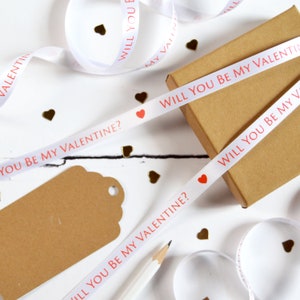 May include: A white satin ribbon featuring the repeating red text WILL YOU BE MY VALENTINE? and small red heart icons. The ribbon is draped over a brown cardboard gift box and accompanied by a brown paper gift tag and a white pencil.