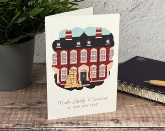 Könnte beinhalten: Eine weiße Grußkarte mit einer Illustration eines roten Backsteinhauses und einem Stapel Kartons mit der Aufschrift fragile davor. Der Text Make Lovely Memories IN YOUR NEW HOME steht in dunkler Schreib- und serifenloser Schrift unten.