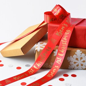 May include: A red satin ribbon featuring gold foil text that reads Ben's 1st Christmas, interspersed with small gold gift box icons. The ribbon is draped over several gift boxes, including one wrapped in red paper and one in brown kraft paper.