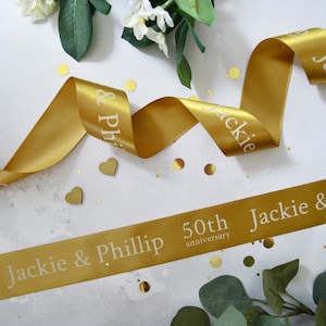 May include: A gold satin ribbon featuring white printed text that reads Jackie & Phillip 50th anniversary. The ribbon is displayed in a flowing, curved arrangement with small gold heart-shaped accents and scattered gold confetti pieces.